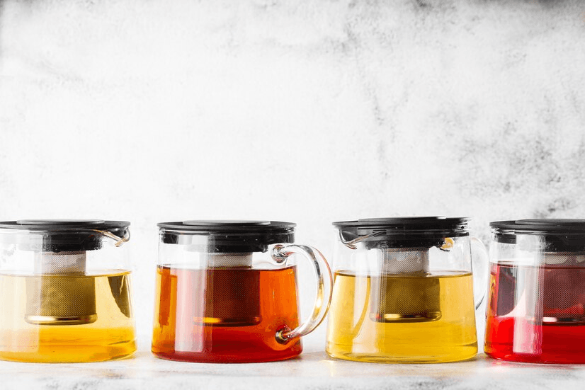 A row of four glass teapots with infusers, showcasing different types of tea in vibrant shades of yellow, amber, and red, placed against a light background.