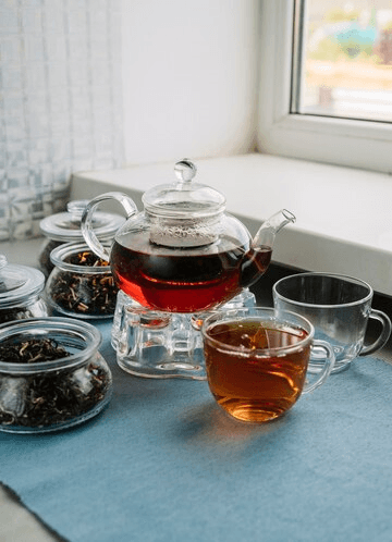 A glass teapot filled with brewed tea placed on a stand with a cup of tea beside it, surrounded by jars of loose tea leaves on a light blue tablecloth by a window.