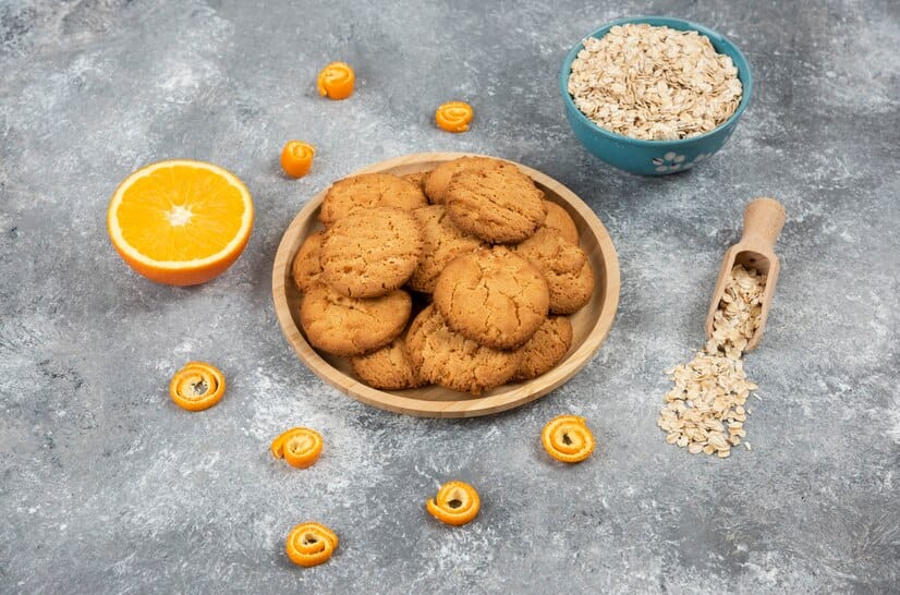 Homemade oatmeal cookies on a wooden plate with oats, orange slices, and orange zest on a gray background