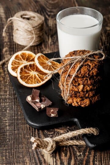 A rustic arrangement of oatmeal cookies tied with twine, accompanied by dried orange slices, chocolate pieces, and a glass of milk on a wooden table.