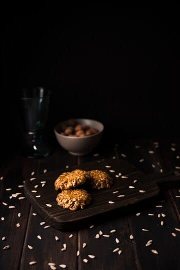 Rustic oatmeal cookies on a wooden board, with scattered oats, a bowl of hazelnuts, and a glass in the background on a dark wooden table.