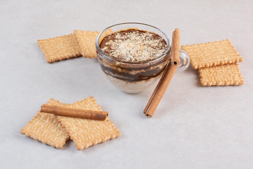 Glass cup filled with layered oatmeal cookie shot topped with crushed cookies and cinnamon, surrounded by biscuits and cinnamon sticks