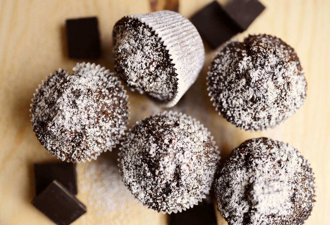 Chocolate protein muffins dusted with powdered sugar, surrounded by dark chocolate chunks, on a wooden surface."