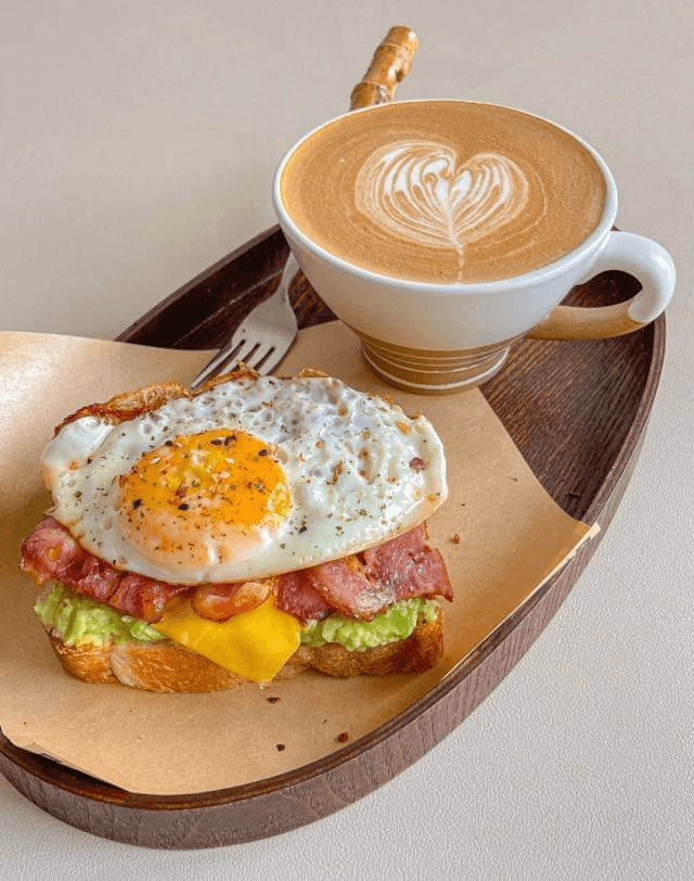 A delicious breakfast of avocado toast topped with a sunny-side-up egg, bacon, cheese, and a latte with heart-shaped foam art on a wooden tray