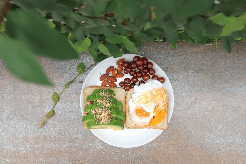 A healthy breakfast plate featuring avocado toast, poached eggs on whole-grain toast, almonds, and fresh red grapes.