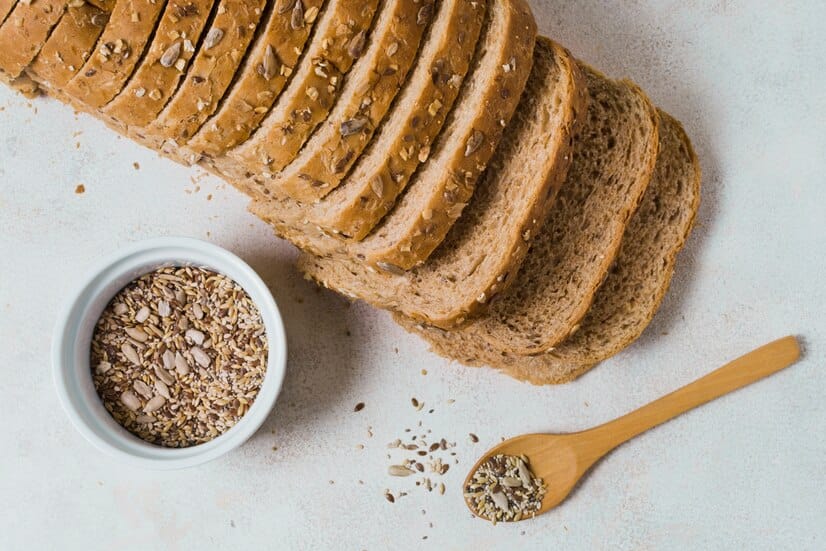 Sliced whole-grain bread topped with seeds, placed on a white surface with a bowl of mixed seeds and a wooden spoon.