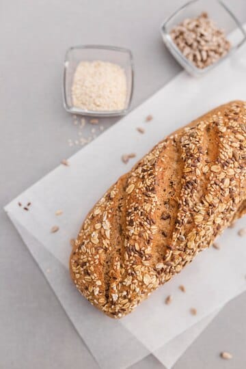 A loaf of seeded bread with a golden crust, placed on parchment paper, surrounded by small bowls of sesame seeds and sunflower seeds