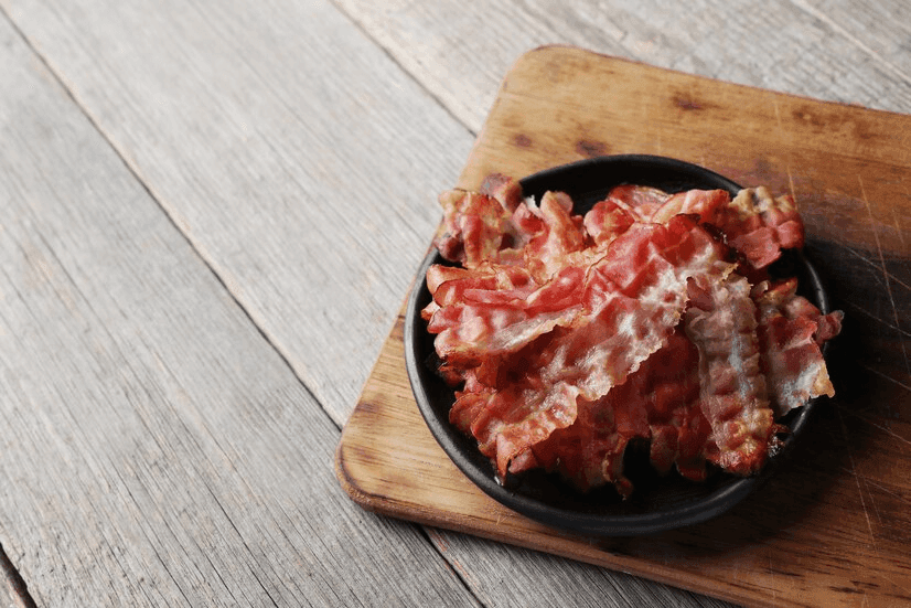 Cooked slices of crispy beef bacon on a wooden cutting board with herbs and spices.