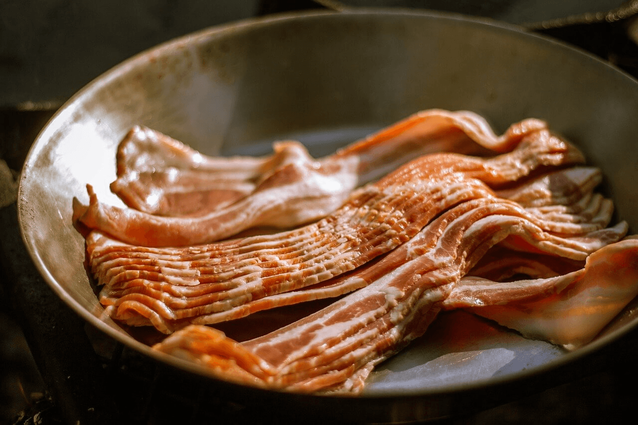 Uncooked strips of beef bacon sizzling in a stainless steel pan under warm lighting.