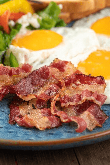 Crispy beef bacon served with sunny-side-up eggs, fresh salad, and toast on a blue plate