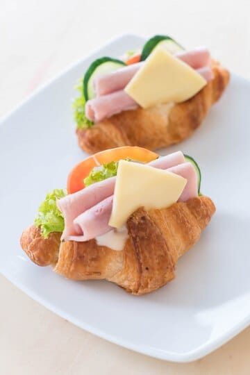 Mini croissants topped with ham, cheese, lettuce, tomato, and cucumber on a white plate