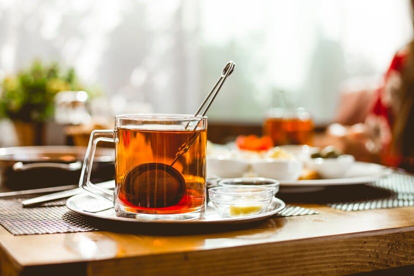 A glass cup of tea with a tea strainer placed on a tray, accompanied by small bowls of honey and lemon, set on a wooden table with a sunny breakfast background.