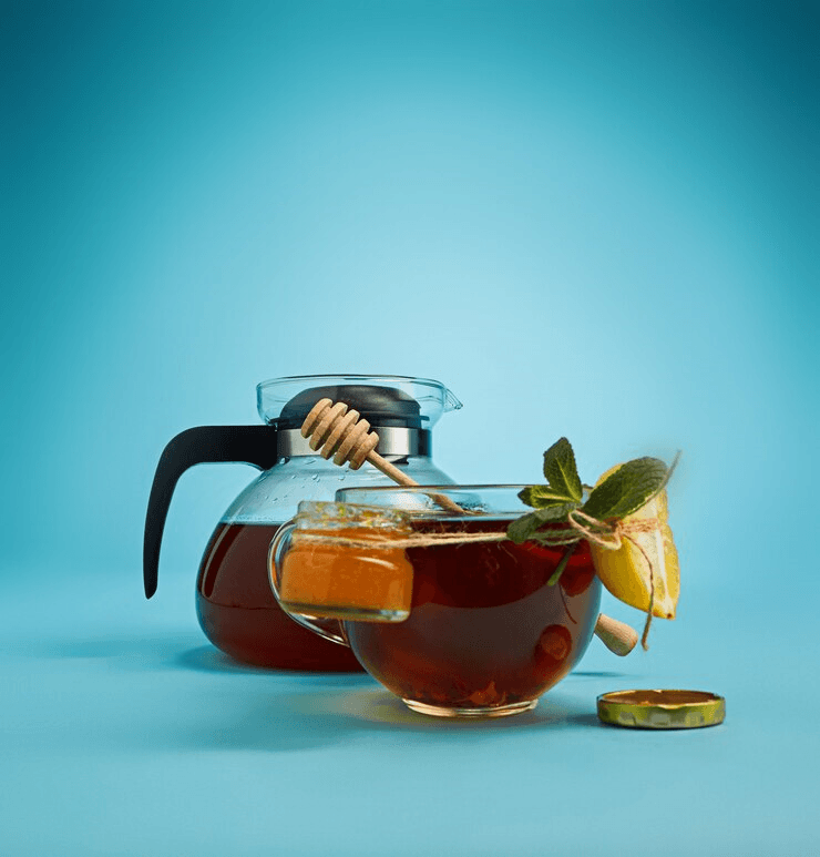 A glass teapot filled with herbal tea alongside a glass cup garnished with mint leaves, lemon slices, and a honey dipper, set against a vibrant blue background.