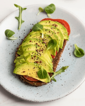 A plate of vegan avocado toast topped with sliced avocado, tomato, and fresh greens on whole-grain bread."