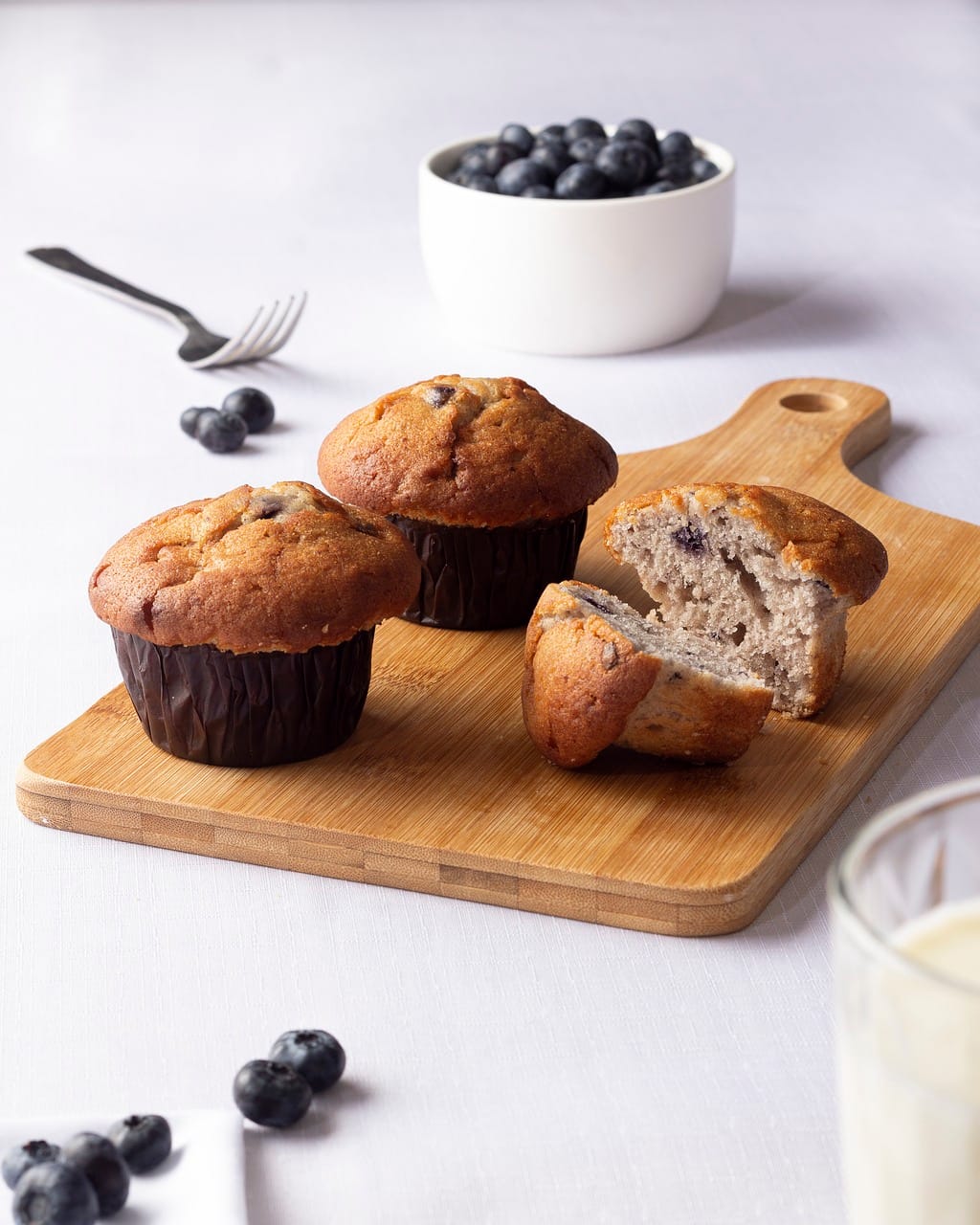 Golden-brown blueberry muffins placed on a wooden cutting board with fresh blueberries in a bowl and scattered on the table.