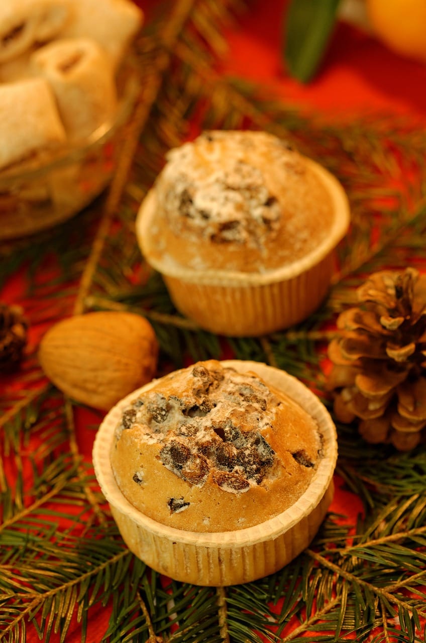 Festive muffins with chocolate chips, lightly dusted with powdered sugar, surrounded by pine branches, walnuts, and a pinecone on a holiday-themed table