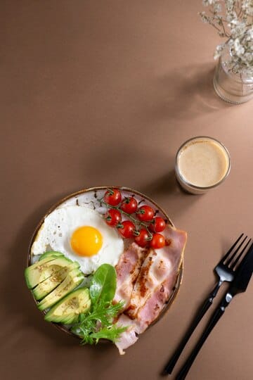 A healthy breakfast plate featuring a fried egg, bacon, sliced avocado, cherry tomatoes, and greens, served with a glass of coffee.