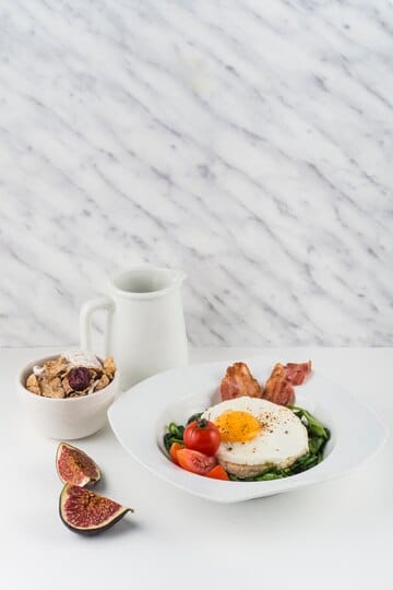 A healthy breakfast plate featuring a fried egg on a bed of spinach, crispy bacon, cherry tomatoes, and figs, served alongside a bowl of granola and a milk jug.