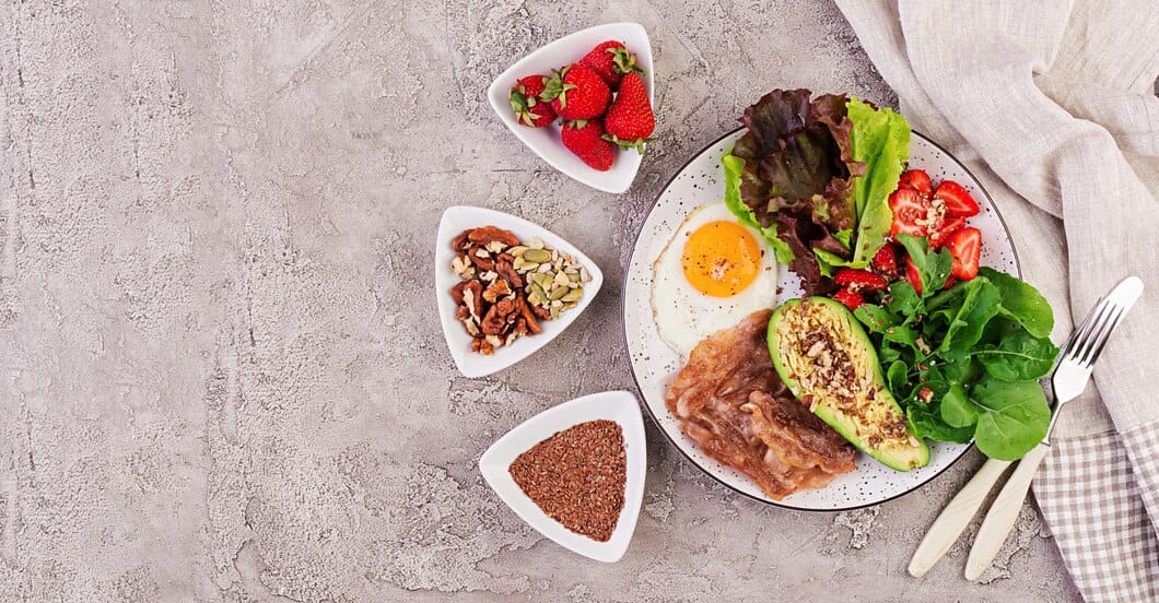 A healthy breakfast plate with a fried egg, bacon, avocado topped with seeds, fresh arugula, lettuce, strawberries, and small bowls of nuts, flaxseeds, and fresh strawberries.