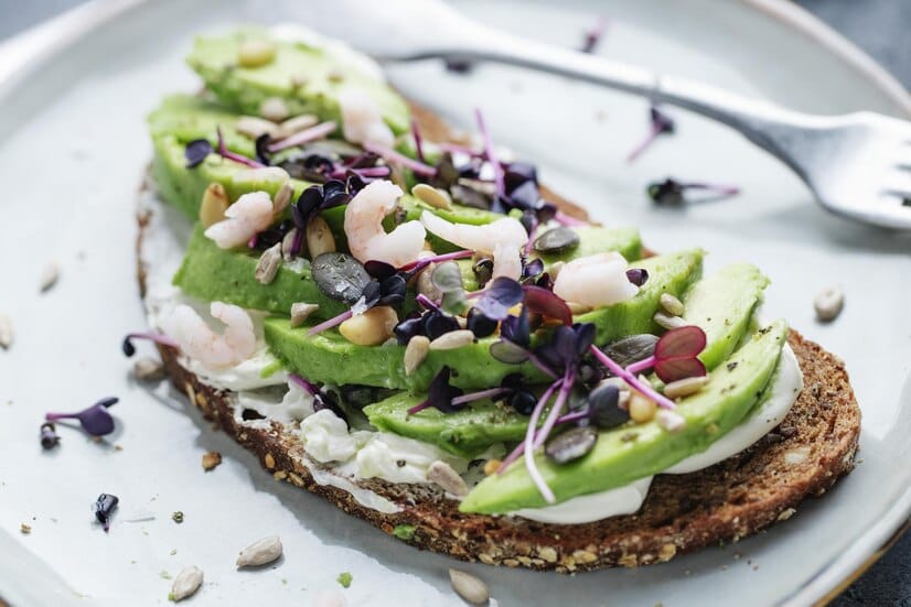 Avocado toast with fresh shrimp, microgreens, and seeds on multigrain bread served on a plate.