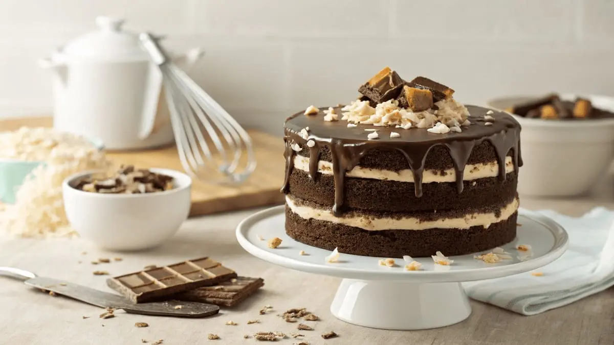 A decadent chocolate cake with rich layers of moist chocolate sponge, creamy coconut filling, and glossy ganache dripping down the sides, topped with Mounds candy pieces and toasted coconut, presented on a white cake stand with baking tools in the background